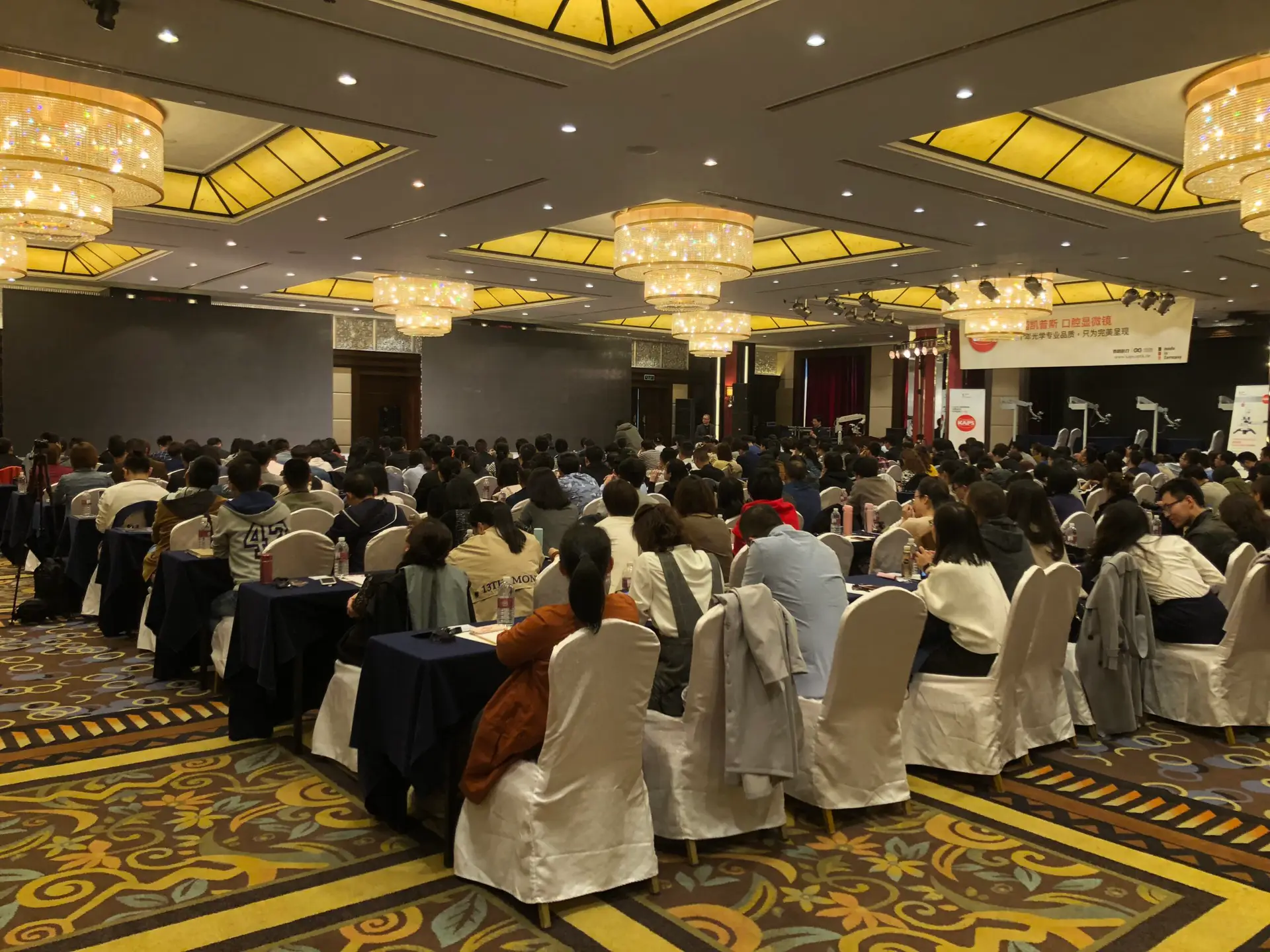 Indoor event with many seated professionals. Sign mentions "Karl Kaps" and "Microscopes made in Germany".