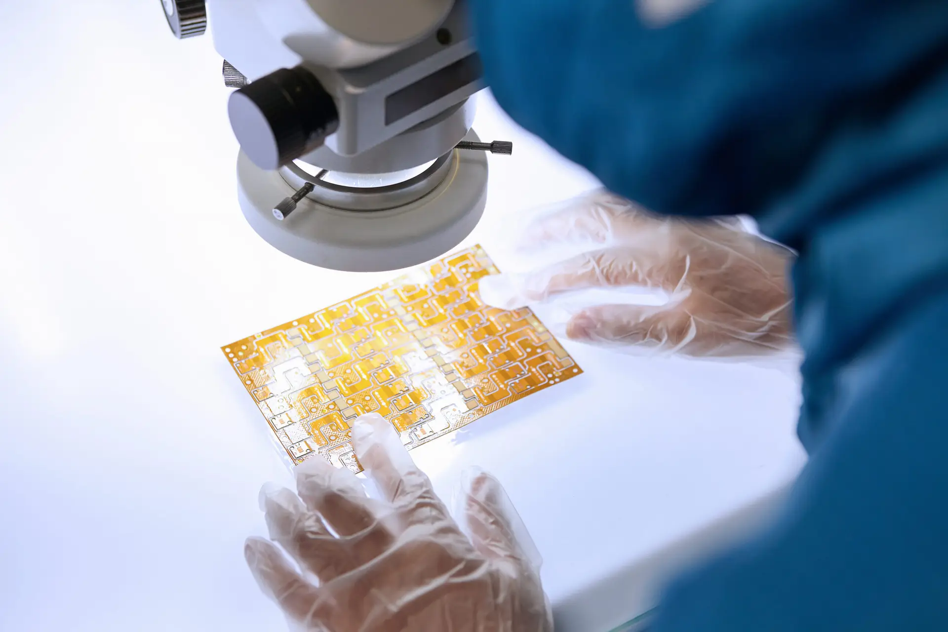Over the shoulder view of female worker using microscope to examine flex circuit in flexible electronics factory clean room